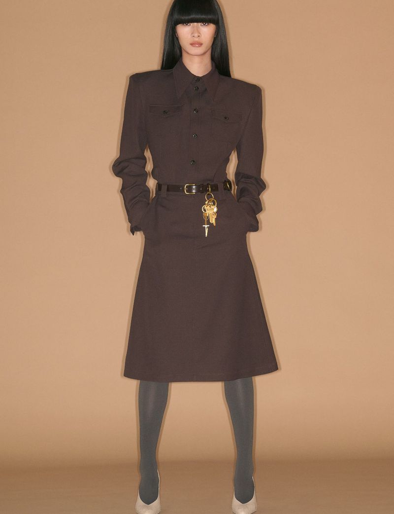 Model standing in a brown belted dress against a plain tan backdrop for System Collections Autumn Winter 2026-2027.