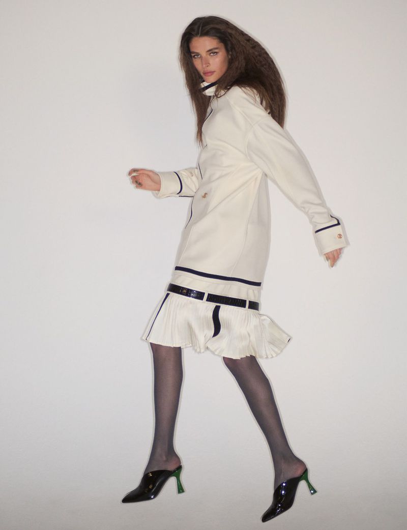 Model wearing an off-white coat with dark trim and dark heels against a neutral background.