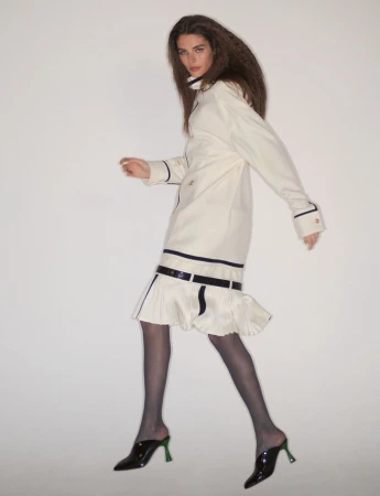 Model wearing an off-white coat with dark trim and dark heels against a neutral background.