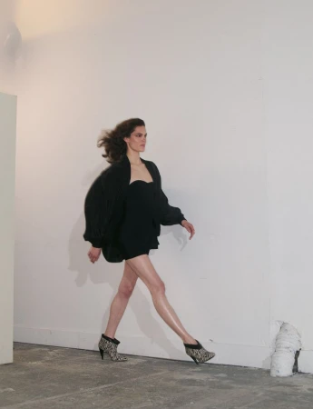 Model walking in a black strapless mini dress and dark oversized jacket against a plain white studio backdrop.
