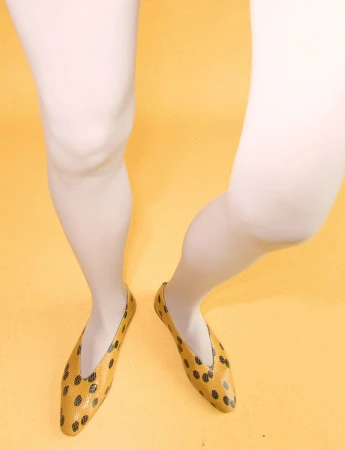 Close-up of legs in white tights wearing yellow patterned flats against a solid yellow studio backdrop.