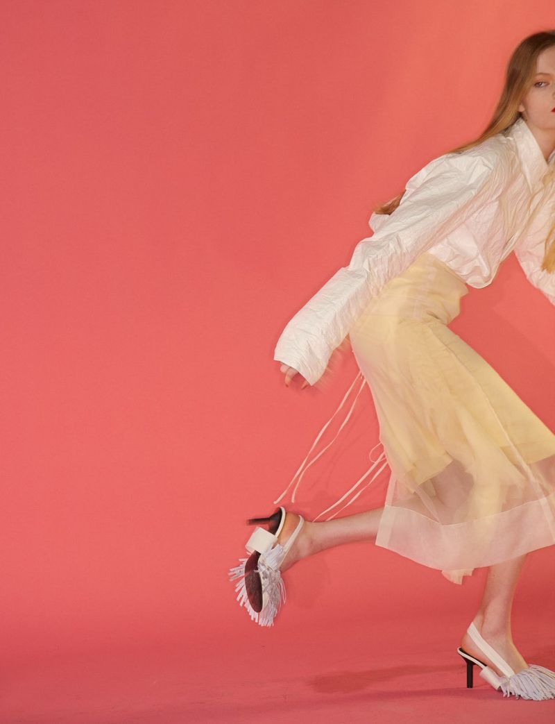 Model wearing a white blouse and yellow sheer skirt with fringed slingback heels against a solid coral background.