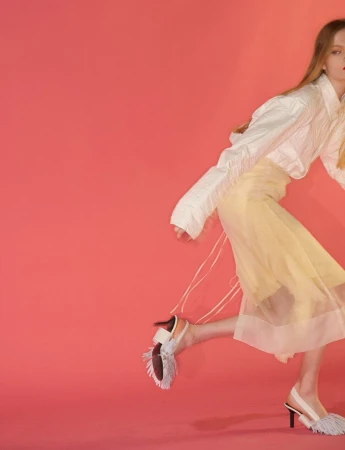 Model wearing a white blouse and yellow sheer skirt with fringed slingback heels against a solid coral background.