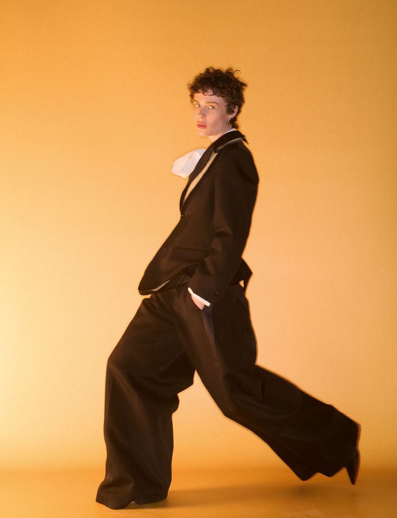 Model striding in a dark suit with wide-leg trousers against a warm yellow studio background for System Collections
