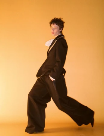 Model striding in a dark suit with wide-leg trousers against a warm yellow studio background for System Collections