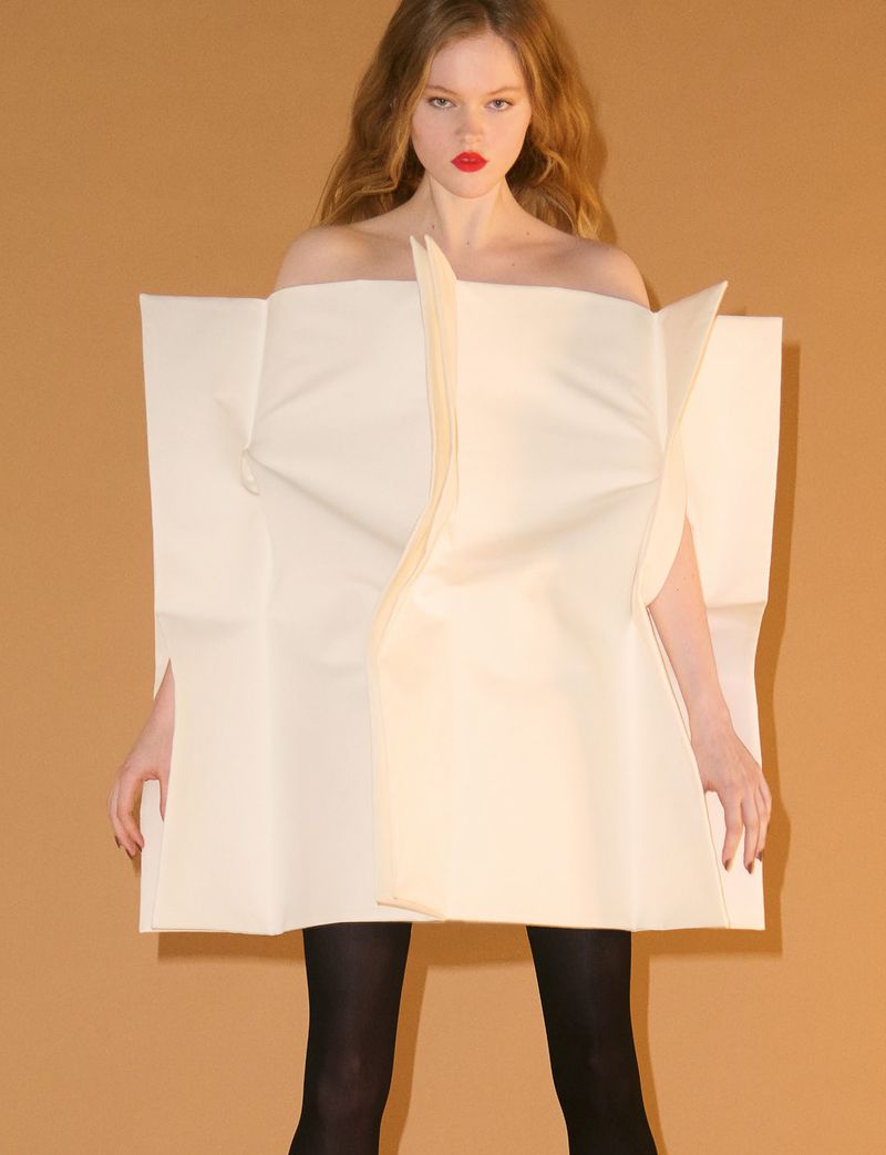 Model wearing a structured cream geometric tunic and dark tights against a warm tan background.