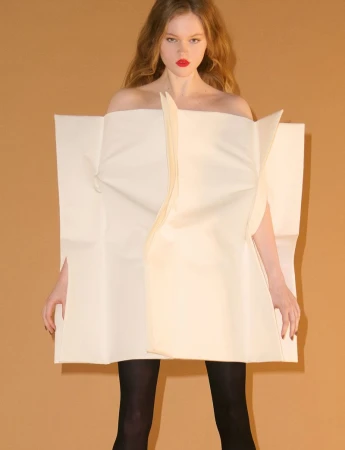 Model wearing a structured cream geometric tunic and dark tights against a warm tan background.