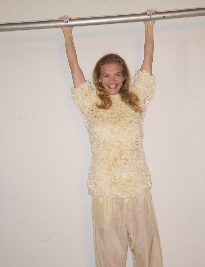 Smiling model hanging from a metal bar in a textured cream top and pale trousers against a plain white background