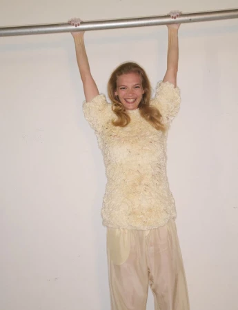 Smiling model hanging from a metal bar in a textured cream top and pale trousers against a plain white background