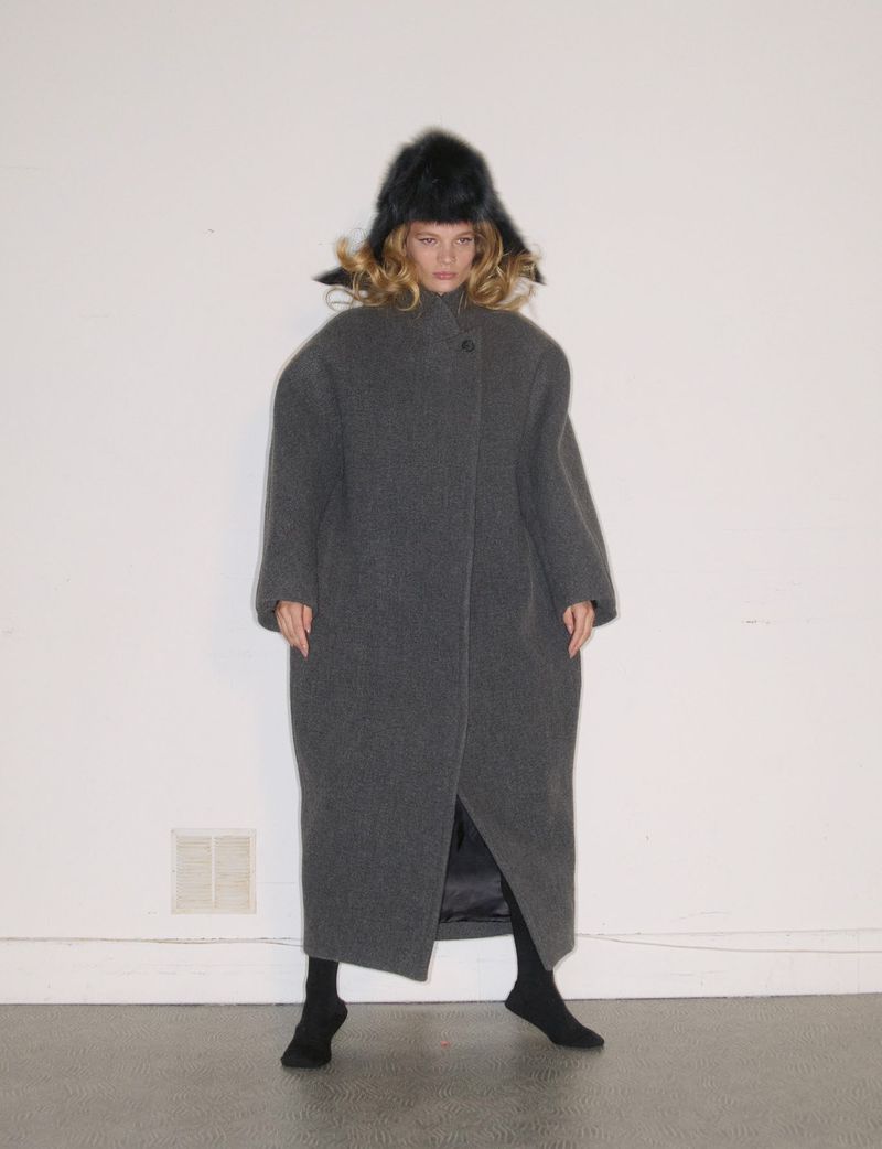 Model in a long grey wool coat and black fur hat standing against a plain wall