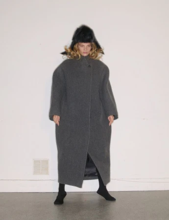 Model in a long grey wool coat and black fur hat standing against a plain wall