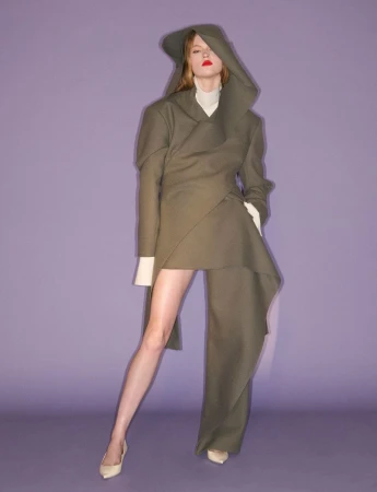 Model wearing an asymmetric hooded olive coat and pale pumps against a solid purple background.