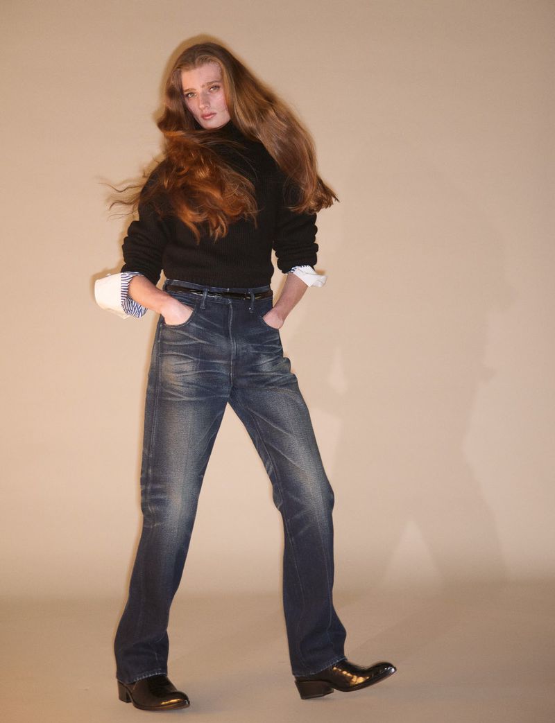 Model with long brown hair wearing a black sweater and dark jeans against a neutral studio backdrop