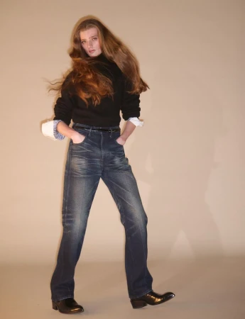 Model with long brown hair wearing a black sweater and dark jeans against a neutral studio backdrop