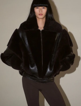 Model wearing a dark shearling jacket and brown trousers against a neutral studio background.