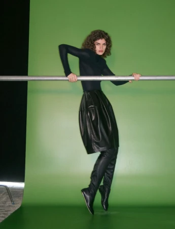 Model in black top and leather skirt leaning on a horizontal metal bar against a bright green backdrop
