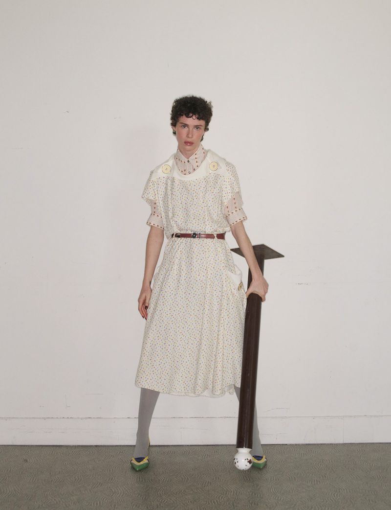 Model in patterned white dress holding a dark pole against a white wall