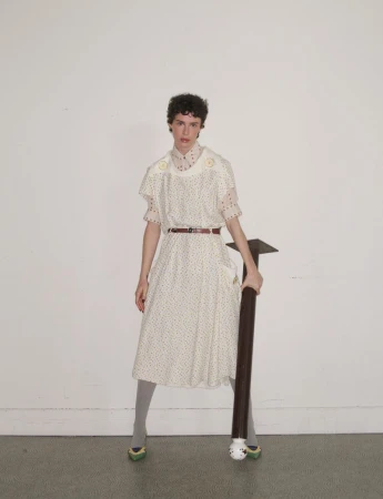 Model in patterned white dress holding a dark pole against a white wall
