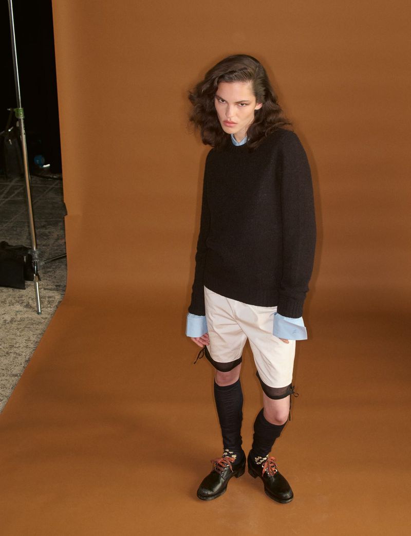 Model wearing a black sweater, light shorts, and dark shoes against a brown seamless studio backdrop.