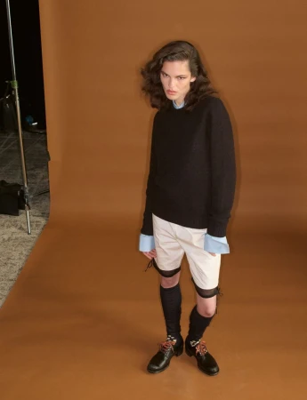 Model wearing a black sweater, light shorts, and dark shoes against a brown seamless studio backdrop.