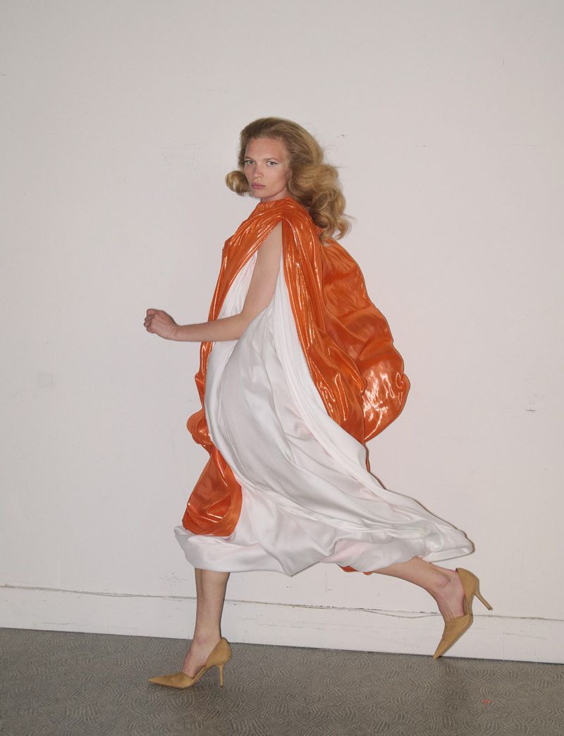 Model wearing a flowing white dress and orange cape running against a plain white background