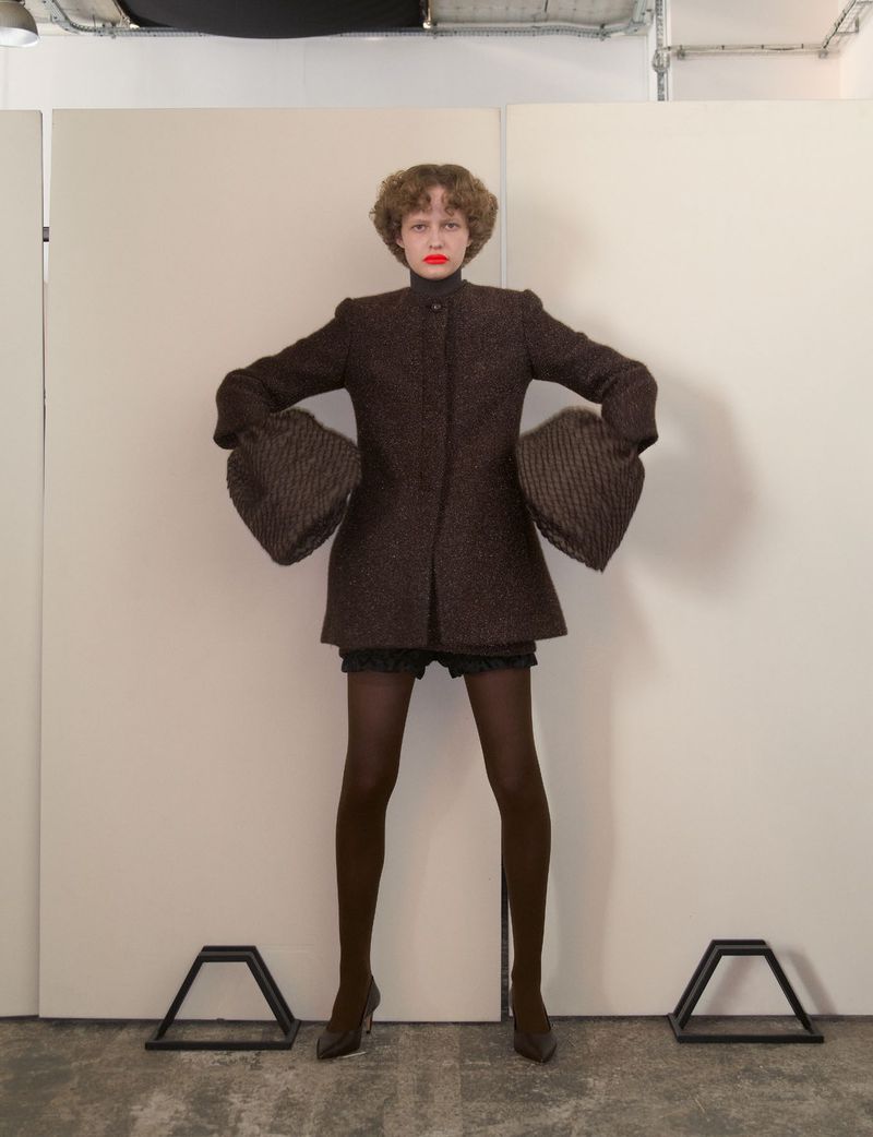 Model wearing dark structured brown jacket with flared cuffs and dark tights against white backdrop