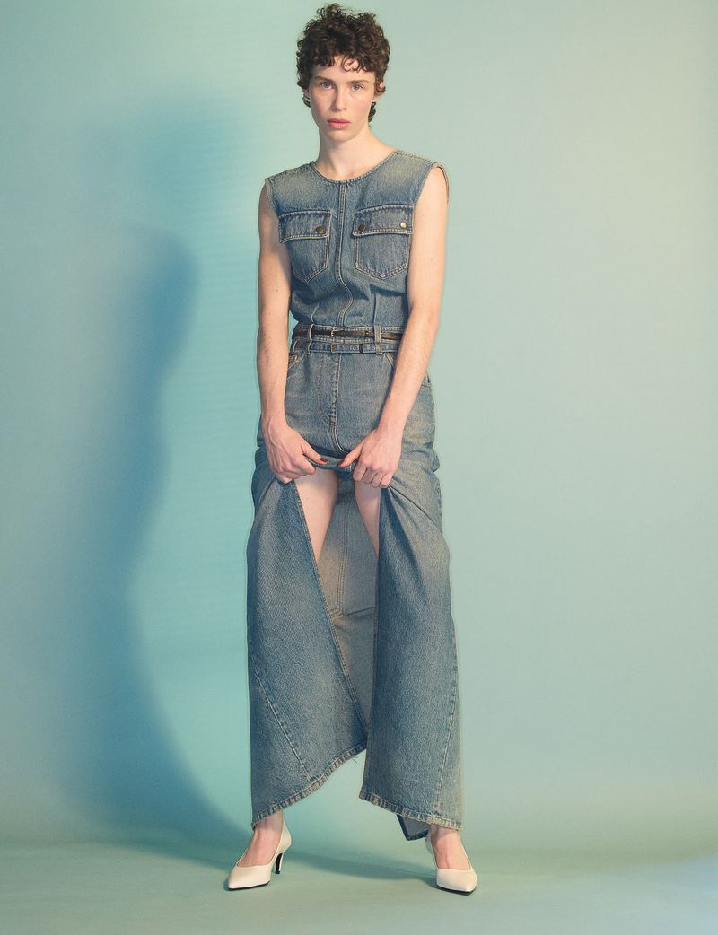 Model standing against a gradient blue background wearing a sleeveless denim dress and white shoes.