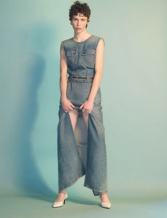 Model standing against a gradient blue background wearing a sleeveless denim dress and white shoes.