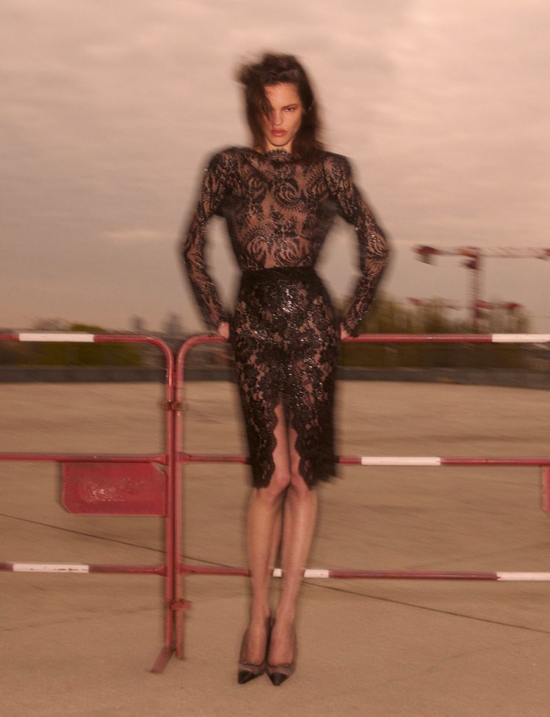 Model wearing a black lace dress leaning against a red and white barrier outdoors under an overcast sky.