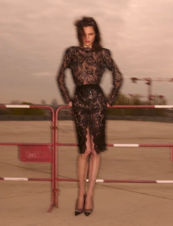 Model wearing a black lace dress leaning against a red and white barrier outdoors under an overcast sky.