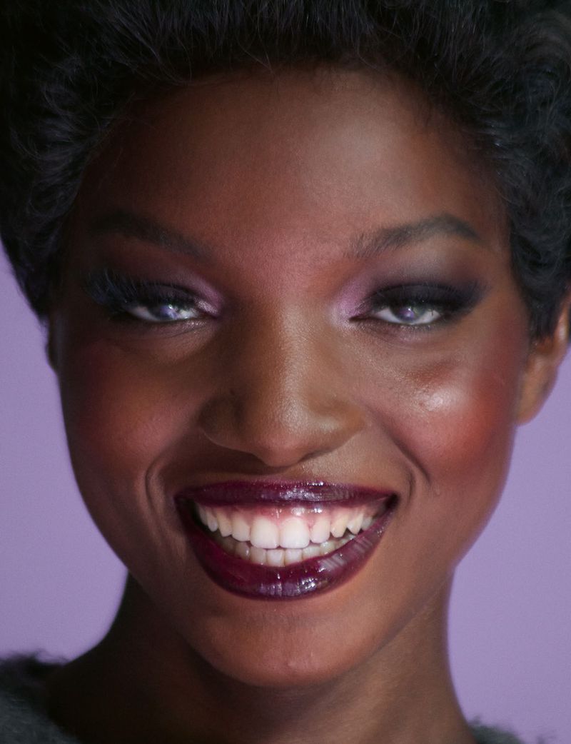 Close-up portrait of a black woman smiling with dark lips and purple eyeshadow against a solid purple backdrop.