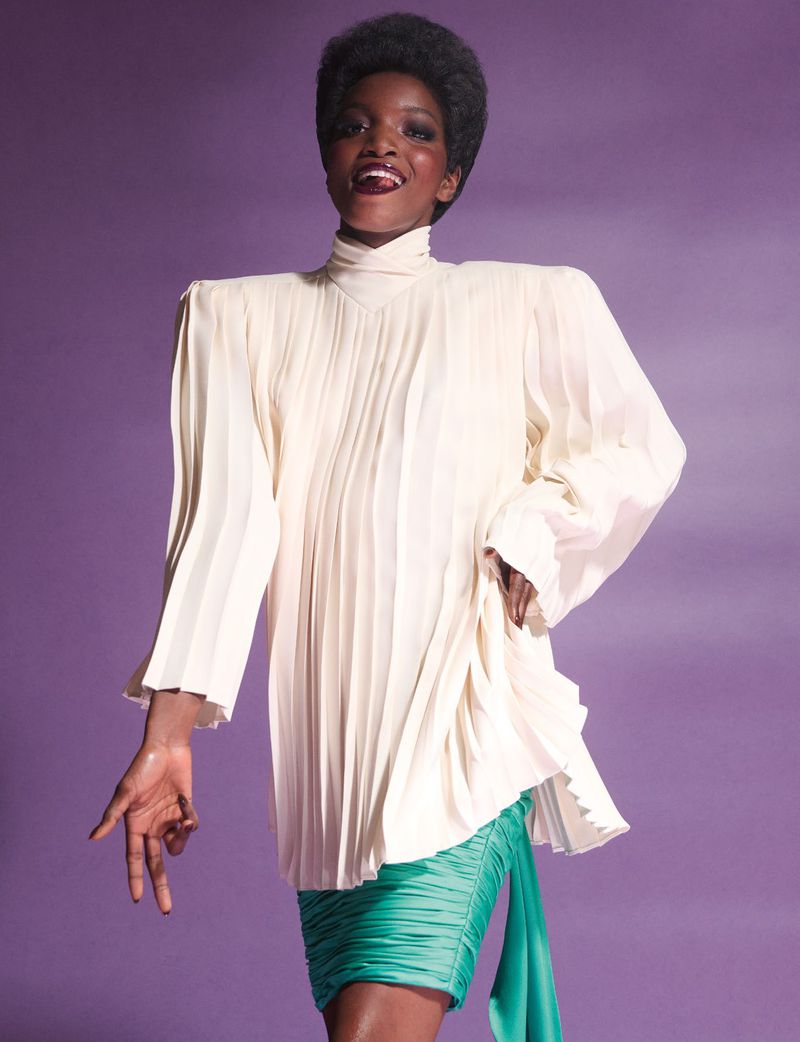 Model in cream pleated blouse and green miniskirt smiling against a purple studio backdrop