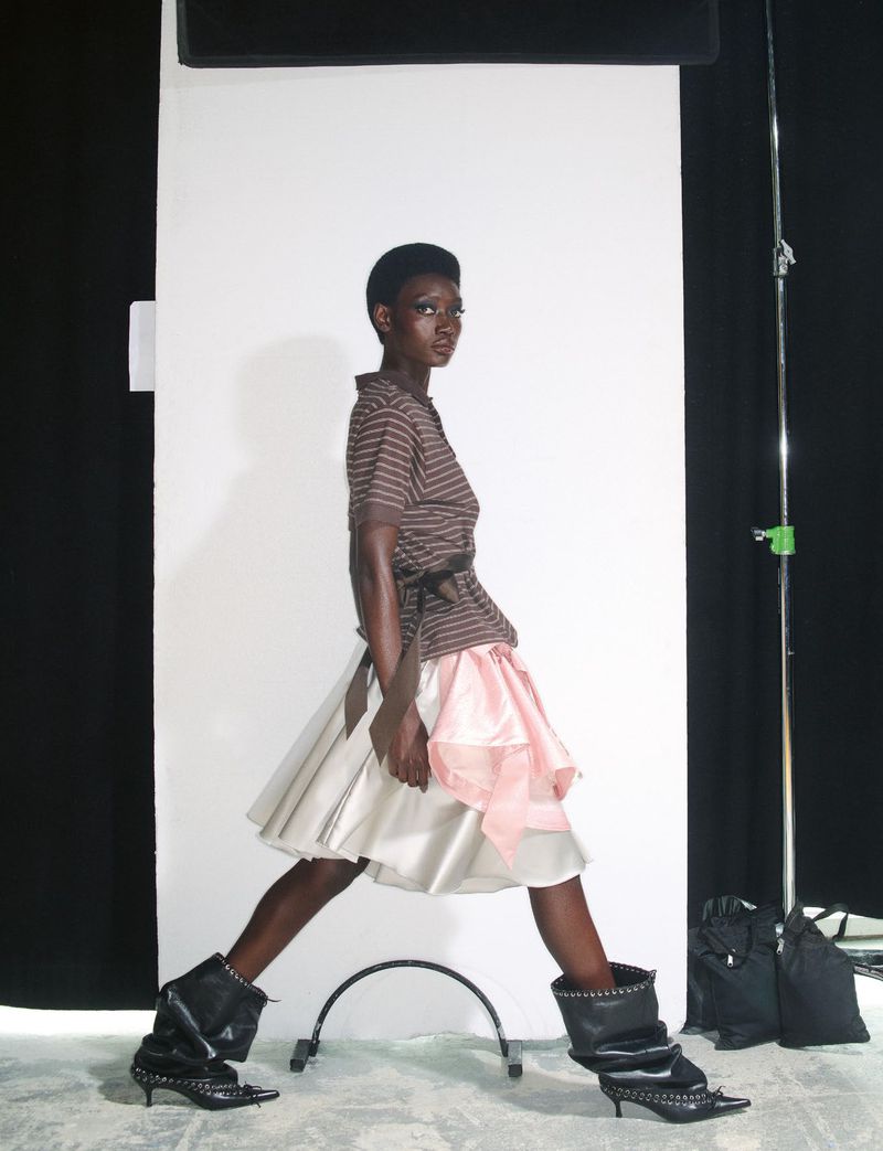 Model in striped brown top and flared skirt walking against a white seamless background with lighting equipment.