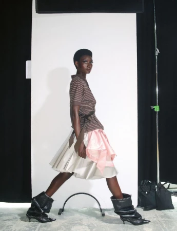 Model in striped brown top and flared skirt walking against a white seamless background with lighting equipment.