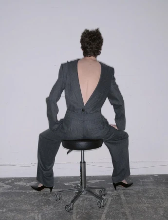 Model seen from behind wearing a charcoal grey suit seated on a rolling office stool against a white wall.