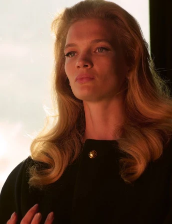 Portrait of a blonde woman wearing a dark coat with gold button against a bright backlit background