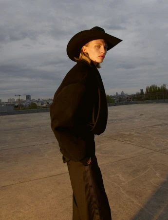 Model wearing a black cowboy hat and dark coat standing on a concrete rooftop under a cloudy sky.