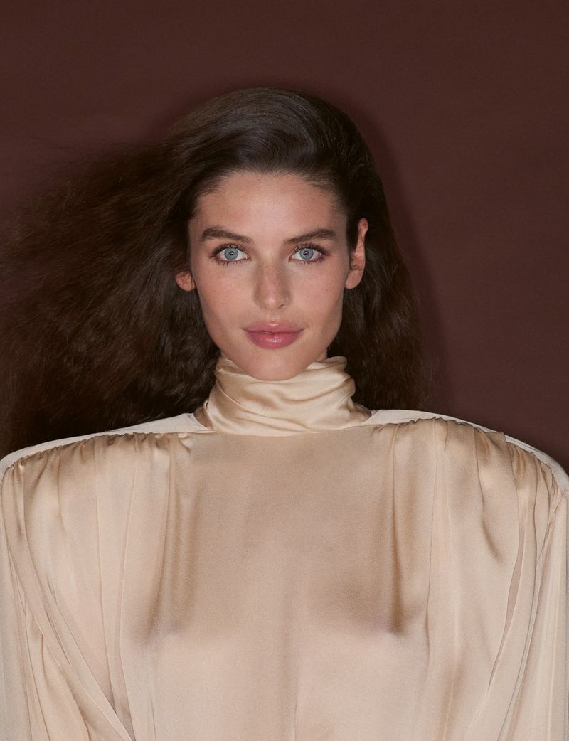Portrait of a woman with dark wavy hair and blue eyes wearing a cream silk turtleneck top against a dark brown background