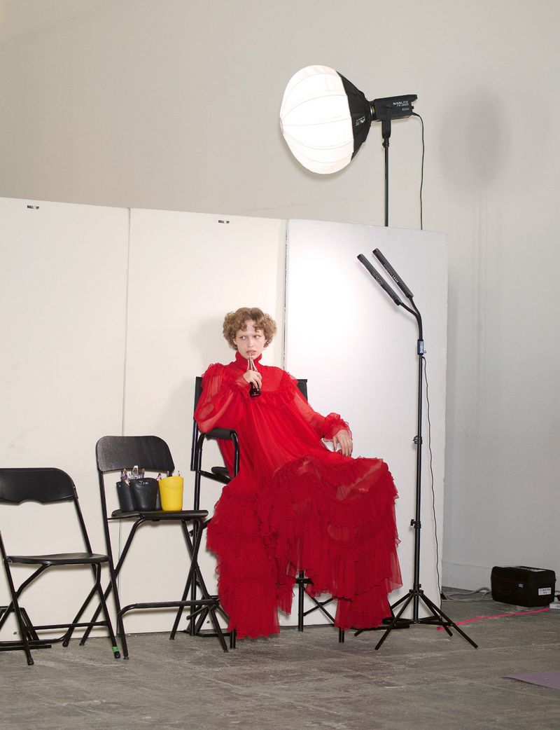 A model in a voluminous red dress seated on a director chair in a studio with lighting equipment