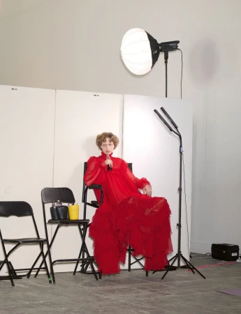 A model in a voluminous red dress seated on a director chair in a studio with lighting equipment