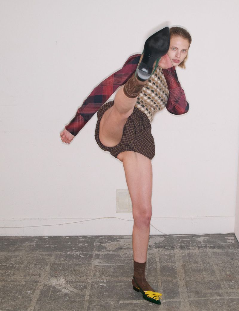 Model in patterned sweater and shorts throwing a high kick against a plain white wall