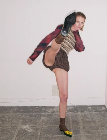 Model in patterned sweater and shorts throwing a high kick against a plain white wall