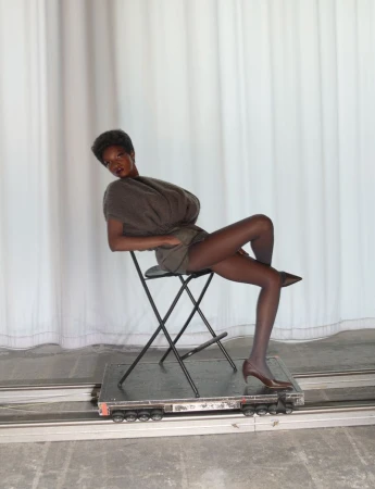 Model seated on folding chair atop wheeled platform against white curtains for System Collections Autumn Winter 2026-2027