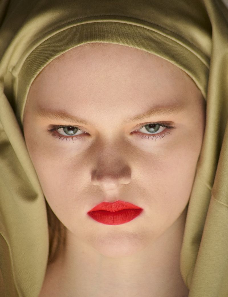 Close-up portrait of a model wearing a golden olive satin hood and vibrant red lipstick by System Collections.
