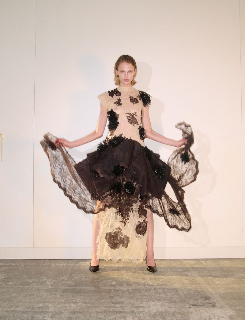 Model standing against a plain wall wearing a cream and dark brown lace dress with floral appliqués, holding the skirt wide.