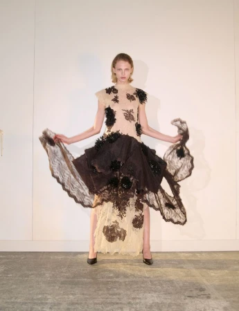 Model standing against a plain wall wearing a cream and dark brown lace dress with floral appliqués, holding the skirt wide.