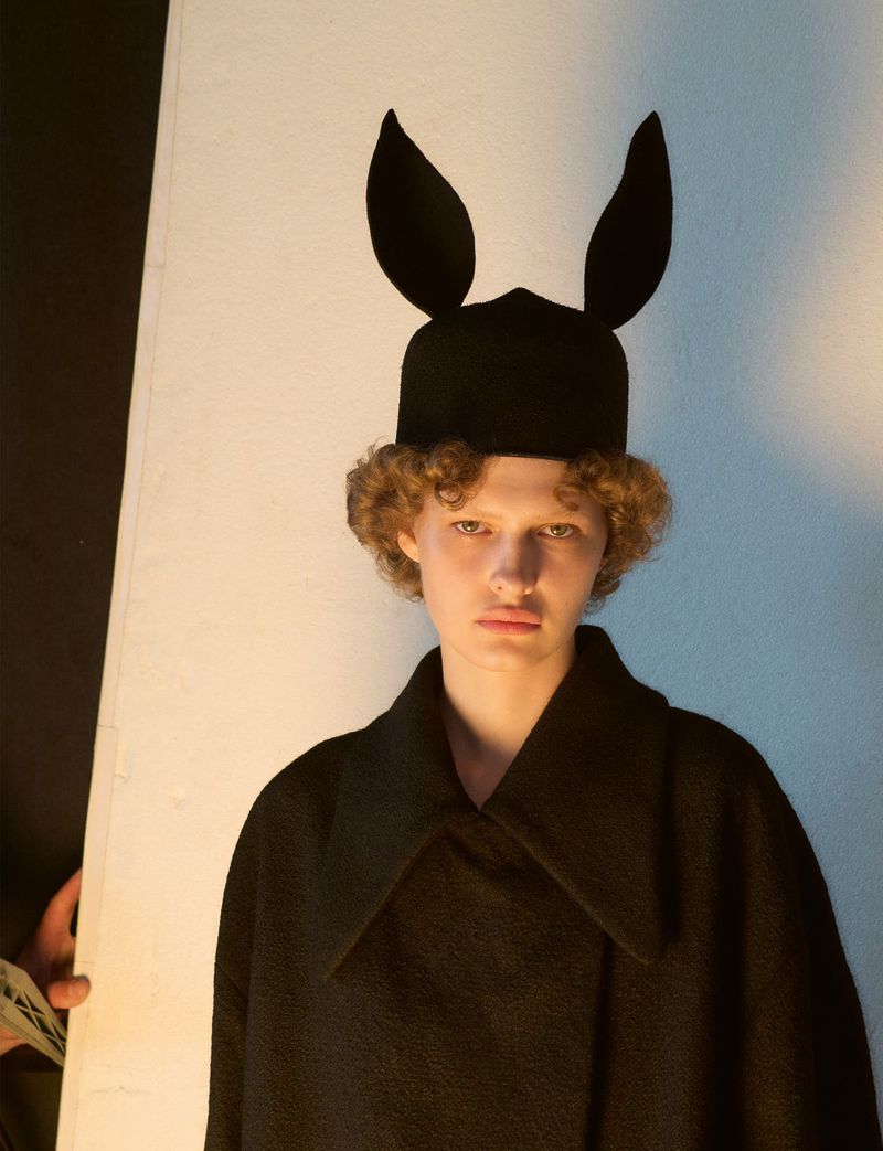 Model wearing a black hat with rabbit ears and a dark coat against a plain background