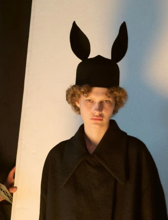 Model wearing a black hat with rabbit ears and a dark coat against a plain background