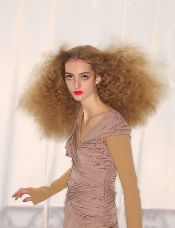 Model with voluminous curly hair and bright red lipstick wearing a ruched mauve dress against a plain backdrop