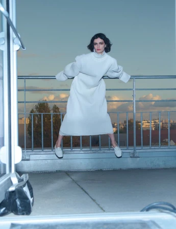 Model wearing a white dress and white shoes leaping in front of a metal railing against a twilight city sky.